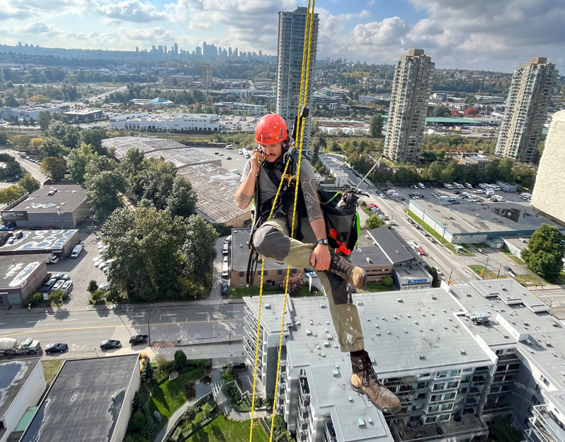 A rope access technician hangs on ropes whilst on a phone call with the city in the background