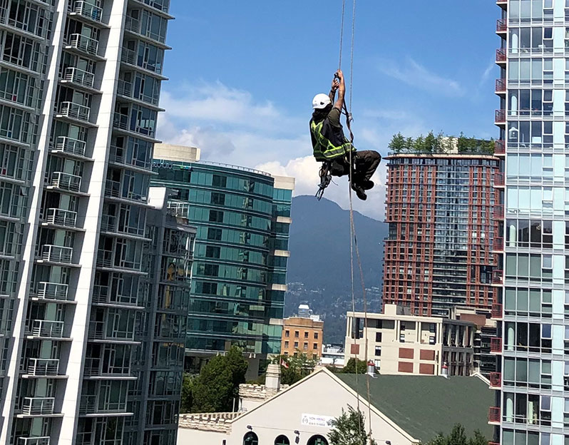 A rope access worker climbs ropes hanging of bc place with Vancouver City in the Background
